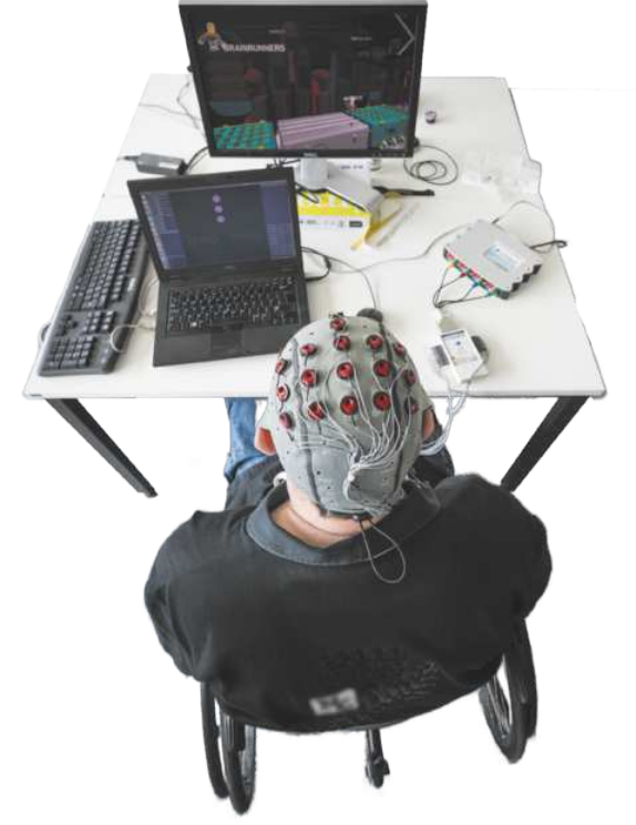 Fotografia de um homem sentado à mesa, usando uma touca com eletrodos conectados a um aparelho ligado a um notebook. Sobre a mesa, também há um teclado e um monitor.