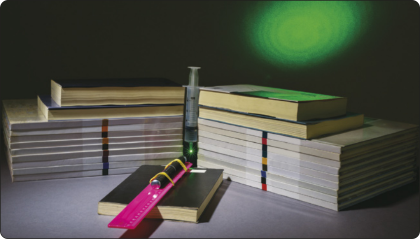 Fotografia do experimento. Em primeiro plano, está o laser preso com elásticos em uma régua apoiada sobre um livro. No meio estão as duas pilhas de livros com seringa no meio. Em segundo plano, está um círculo de luz na parede.