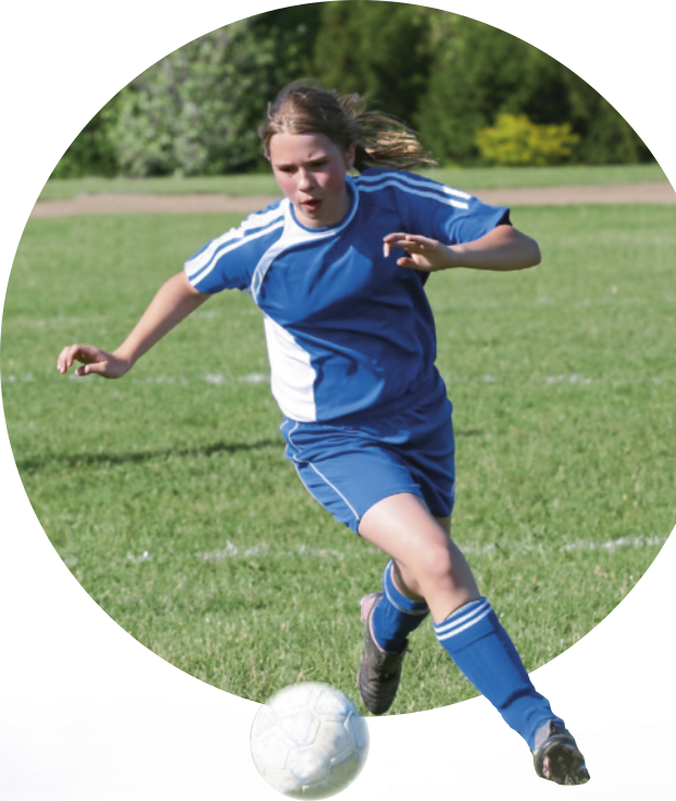 Fotografia de uma mulher de pele clara, cabelos longos, lisos e presos, correndo ao ar livre, prestes a chutar uma bola de futebol. Ela usa uniforme e chuteiras.