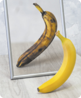 Fotografia de uma banana com a casca sem manchas em frente a um espelho, porém seu reflexo mostra uma banana com a casca repleta de manchas escuras e amassados.
