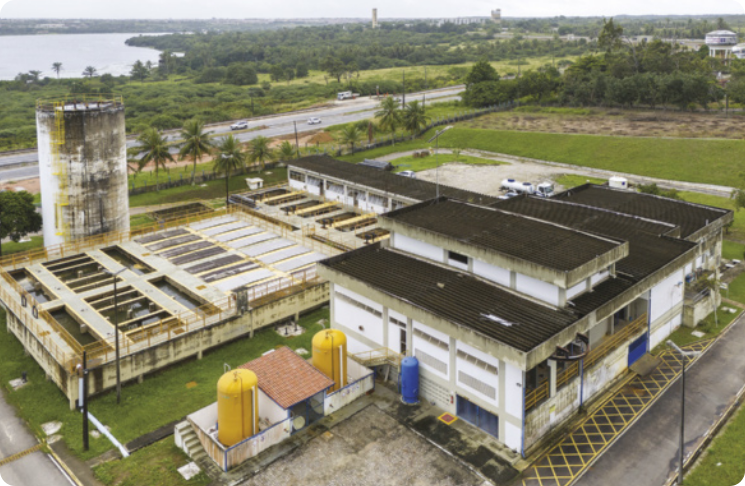 Fotografia aérea de uma estação de tratamento de água. No primeiro plano, há um conjunto de edifícios, estruturas industriais e grandes compartimentos abertos. À esquerda, há uma torre alta e cilíndrica. Ao fundo, vê-se uma estrada com alguns carros passando, vegetação abundante e, mais distante, um corpo d’água.