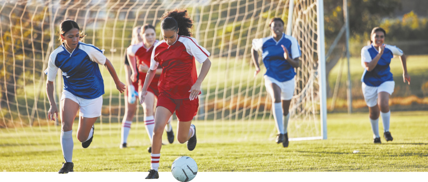 Fotografia de mulheres jovens jogando futebol em um campo ao ar livre. Todas usam uniformes e chuteiras.