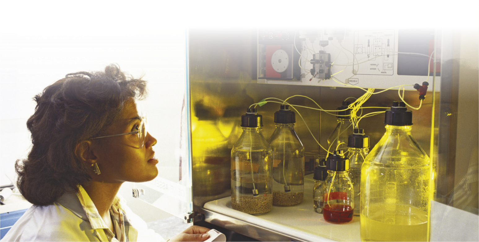 Fotografia de uma mulher negra de óculos observando frascos contendo líquidos diferentes. Das tampas dos frascos saem tubos finos que estão conectados a uma máquina acima deles.