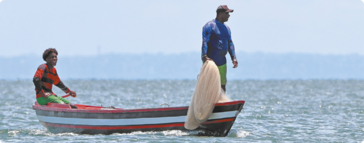 Fotografia de dois homens sobre um barco no mar. Um deles está sentado e o outro está de pé segurando uma rede de pesca.