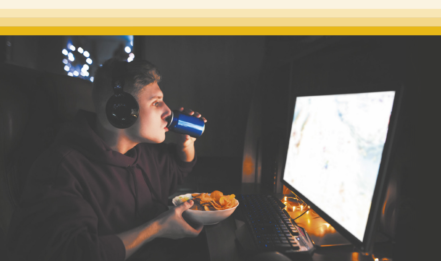 Fotografia de um jovem de cabelos curtos, vestindo um moletom, sentado em frente a um computador, usando fones de ouvido, enquanto bebe refrigerante de uma lata e segura uma tigela de salgadinhos.