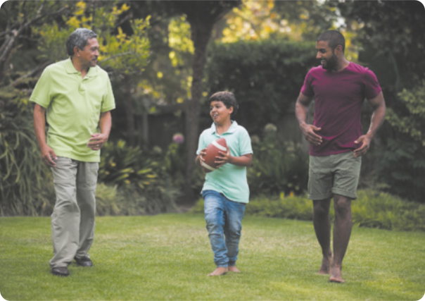Fotografia de dois homens e um menino em um jardim, todos têm pele escura. À esquerda, está um homem de cabelos curtos e grisalhos, veste camisa, calça e sapato. Olha para os outros dois sorrindo. No centro, está o menino que sorri e segura uma bola de futebol americano. Usa camisa e calça. Está com os pés descalços. À direita está um homem de cabelo curto, que sorri enquanto olha para o menino. Usa camiseta e bermuda. Também está com os pés descalços.