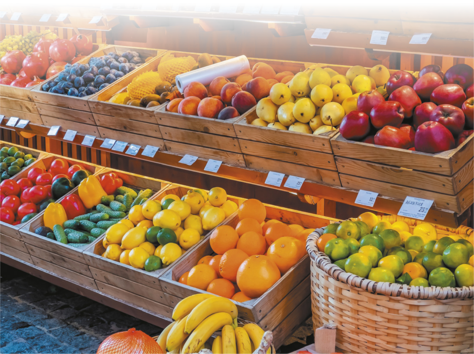 Fotografia de frutas e vegetais dispostos em caixas de madeira. Laranjas estão em um cesto de palha.