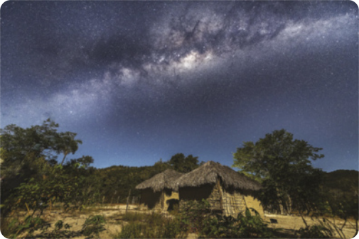 Fotografia ao ar livre de duas casas de pau a pique. No céu, está uma grande faixa luminosa. Existe uma mata densa em segundo plano.