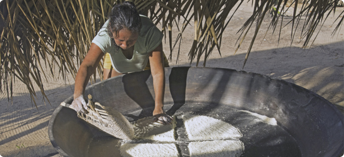 Fotografia de mulher indígena distribuindo a farinha de mandioca sobre uma grande panela de barro ao ar livre. A mulher tem cabelo longo, liso e preso. Veste camiseta e shorts. Usa um utensílio de palha para dar formato ao beiju.