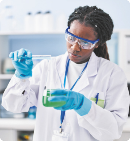 Fotografia de uma mulher negra de tranças com expressão concentrada em um laboratório. Ela usa óculos de proteção, luvas e um jaleco enquanto transfere um líquido de um tubo de ensaio para um frasco.