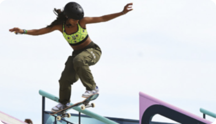 Fotografia de uma mulher negra de cabelos longos fazendo uma manobra com um skate em um corrimão. Ela usa calça, top, tênis e capacete e tem ambos os pés posicionados sobre o skate. Seu tronco está inclinado para frente e tem os braços abertos na altura dos ombros. O centro do skate está apoiado no corrimão.