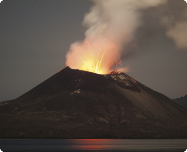 Fotografia de um vulcão em erupção. Existe lava e fumaça saindo de seu topo. Na base dele, existe um corpo de água.