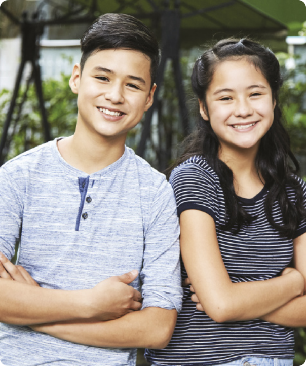 Fotografia de dois adolescentes, um menino e uma menina com traços orientais. O menino tem cabelo curto e liso, a menina tem cabelo longo e liso. Ambos usam camiseta, têm os braços cruzados e sorriem para a foto.