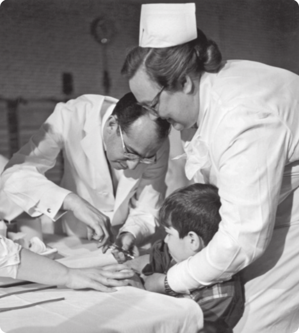 Fotografia em preto e branco de médico aplicando vacina no braço de um menino. Duas enfermeiras estão auxiliando.