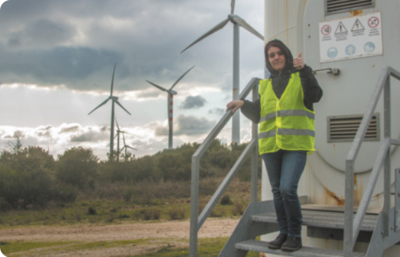 Fotografia de engenheira de energia renovável eólica. Ela está sobre degraus de uma escada ao ar livre, usa colete refletor e ao fundo há torres de energia eólica.