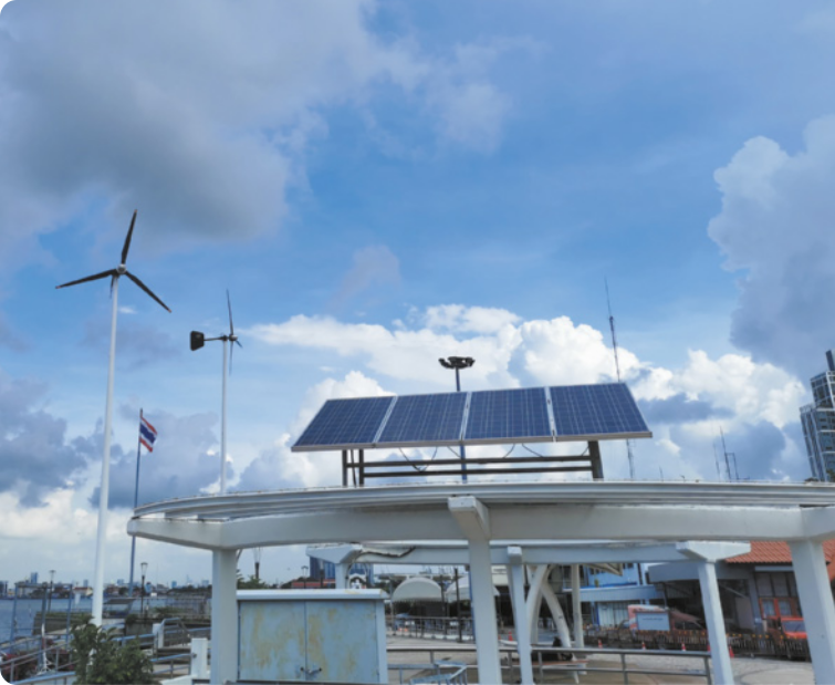 Fotografia de usina mista de geração de energia. Há um painel solar exposto e duas torres eólicas.