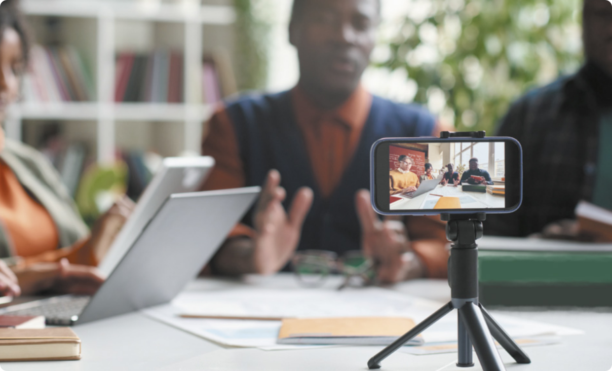 Fotografia de pessoas fazendo reunião, usando notebooks e conversando entre si. Há um celular em tripé gravando a reunião.