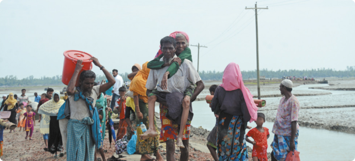 Fotografia de homens e mulheres refugiados andando em solo de barro à margem de um rio. Eles carregam sacolas e baldes. As mulheres usam lenços na cabeça.