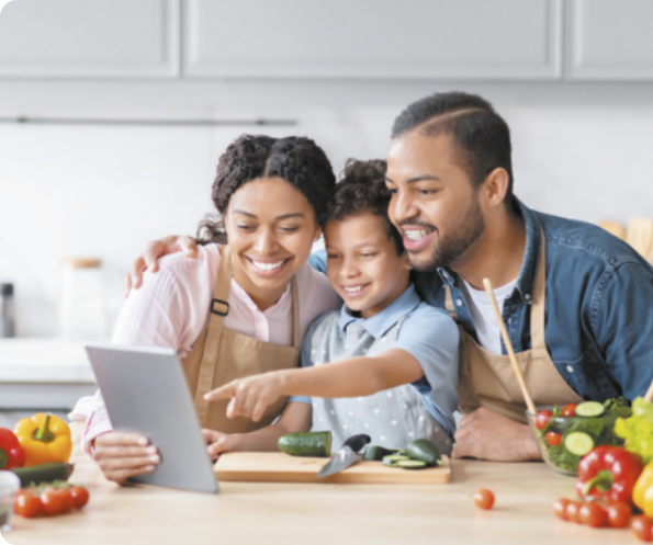 Fotografia de uma família na bancada da cozinha. A mãe segura um tablet, a criança aponta para o tablet e o pai observa.