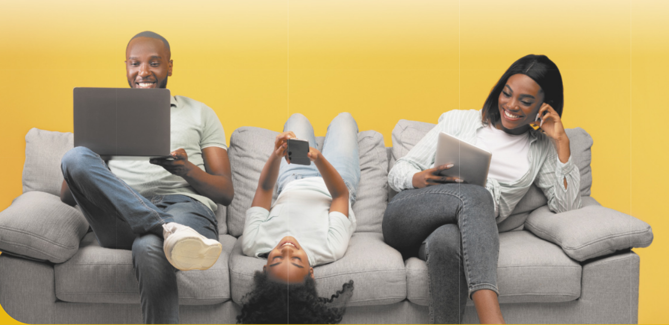 Imagem de homem, criança e mulher sentados no sofá, utilizando notebook, celular e tablet.