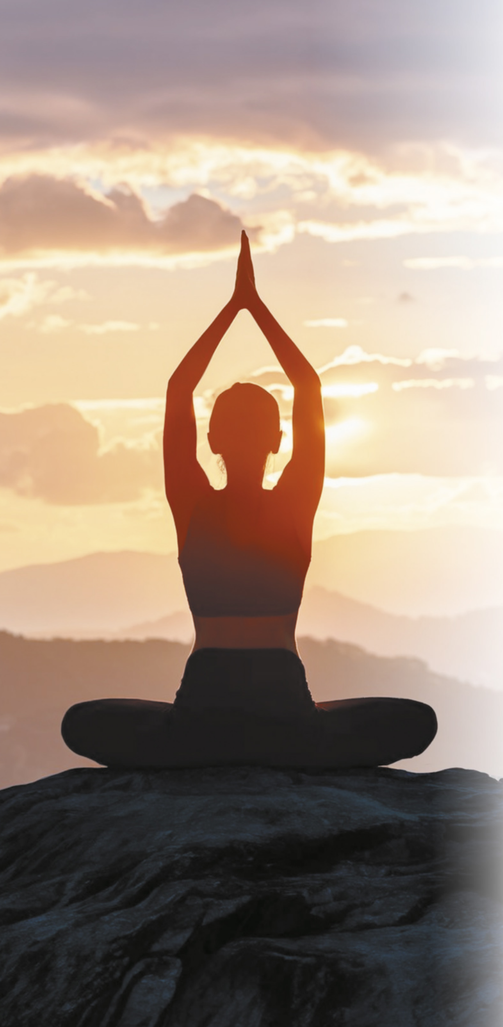 Imagem de mulher em postura de yoga, sentada com as pernas cruzadas e palmas das mãos unidas acima da cabeça. Ela está sentada em uma pedra, no topo de uma montanha.