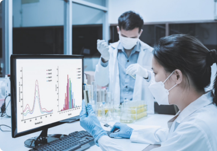 Fotografia de um cientista homem e uma cientista mulher em laboratório. Ele está usando máscara, jaleco, luva e está manipulando amostras em vidraria. Ela está de cabelos presos, usando máscara, jaleco e luva e está analisando dados em gráfico na tela do computador.
