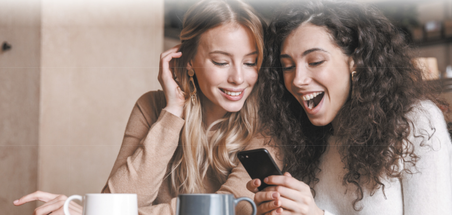 Fotografia de duas mulheres sorrindo e olhando para tela de celular.