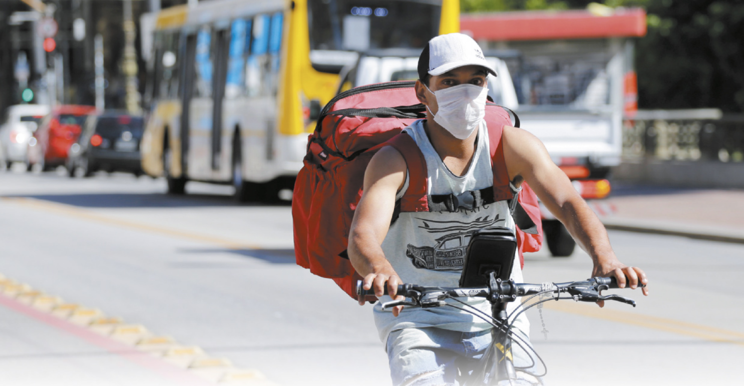 Fotografia de um entregador dirigindo uma bicicleta. Ele usa bolsa de entregas, boné e máscara cobrindo nariz e boca.
