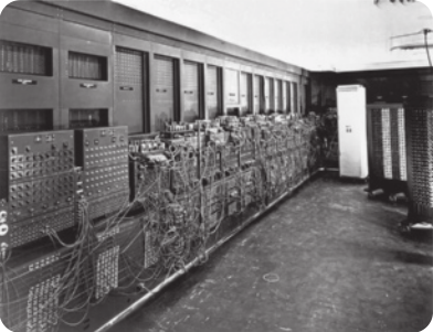 Fotografia em preto e branco de uma grande sala com um computador antigo ocupando toda a extensão de uma das paredes.