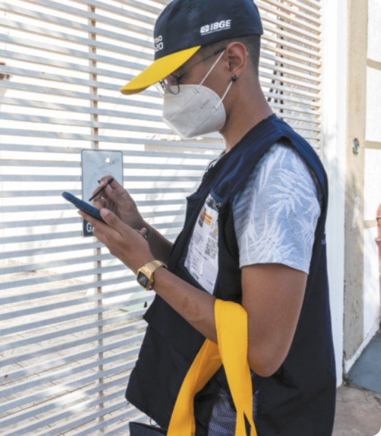 Fotografia de homem usando boné com sigla 'IBGE', colete com sua identificação e máscara cobrindo nariz e boca. Ele está parado em frente ao portão de uma casa e fazendo anotações em um smartphone.