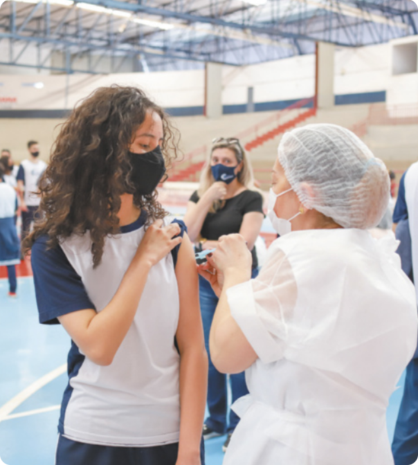Fotografia de enfermeira aplicando vacina no braço de uma menina, que está usando máscara sobre o nariz e boca. Elas estão em um ginásio escolar.