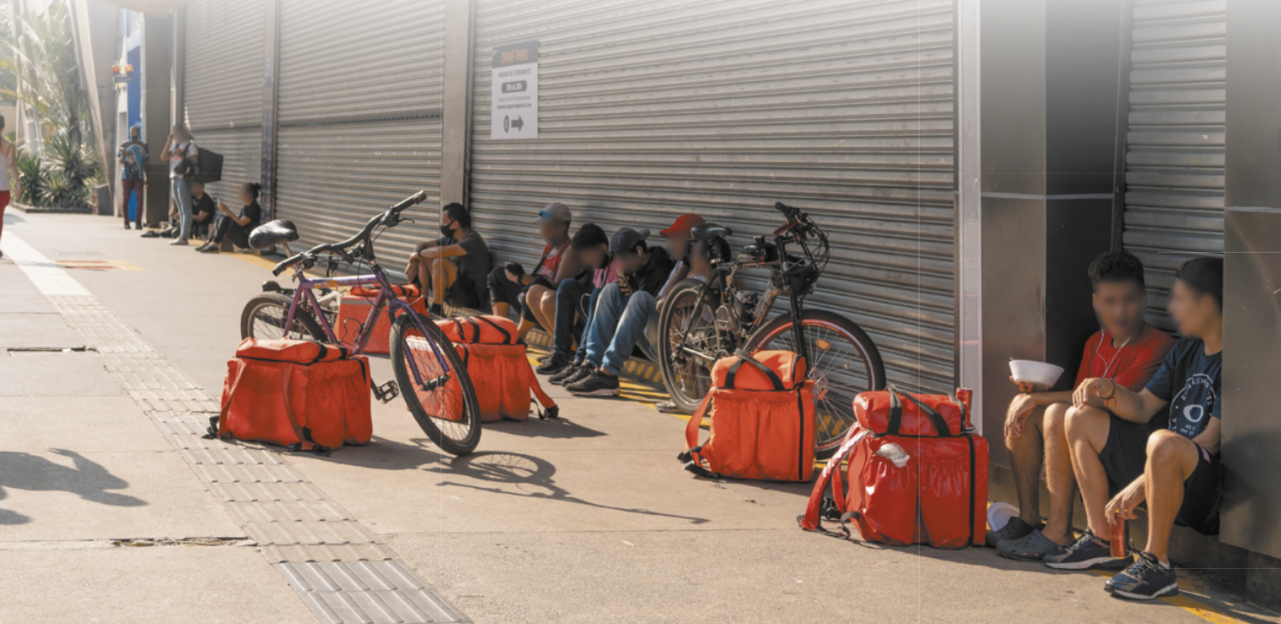 Fotografia de alguns entregadores sentados na calçada em frente a um portão de ferro, com as suas bolsas de entrega e bicicletas sobre a calçada.