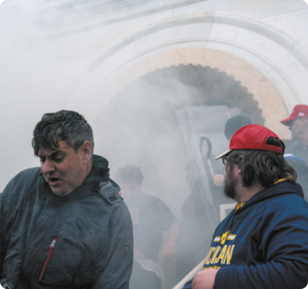 Fotografia de homens manifestantes saindo do prédio do Capitólio. Eles usam casacos e há fumaça.