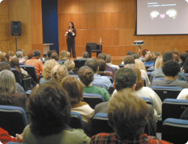 Fotografia de pessoas assistindo à palestra de Suzana Herculano-Houzel em auditório. Ela está sobre palco, falando ao microfone e as pessoas estão ouvindo sentadas em poltronas enfileiradas.