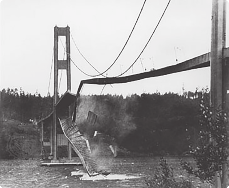 Fotografia em preto e branco de ponte colapsada. Há pedaços da ponte caídos no curso de água abaixo e a paisagem é composta por vegetação.
