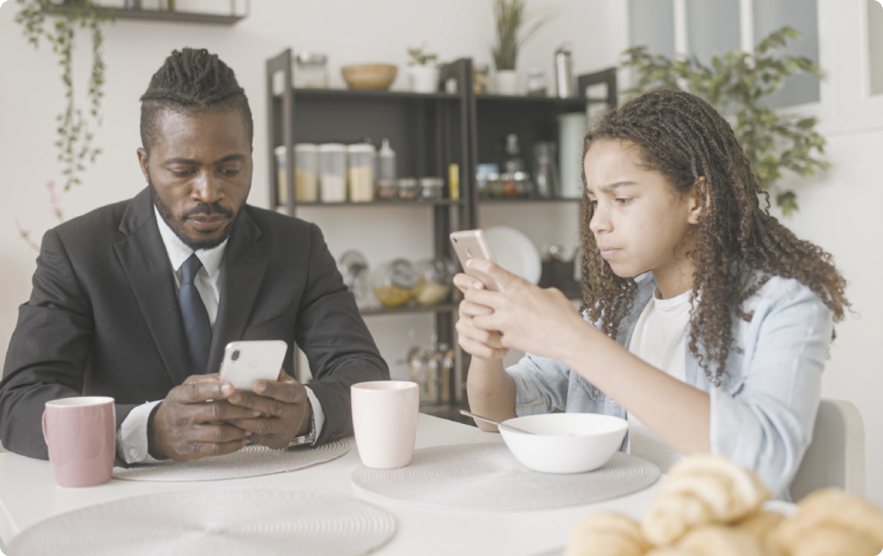 Imagem de um homem e uma menina sentados à mesa de café da manhã, ambos usando seus celulares.