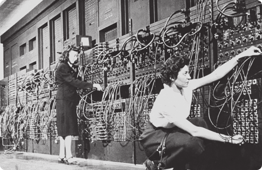 Fotografia em preto e branco de duas mulheres conectando cabos no computador ENIAC. O computador é grande, ocupa toda a altura da sala em que está e possui muitos cabos e botões.