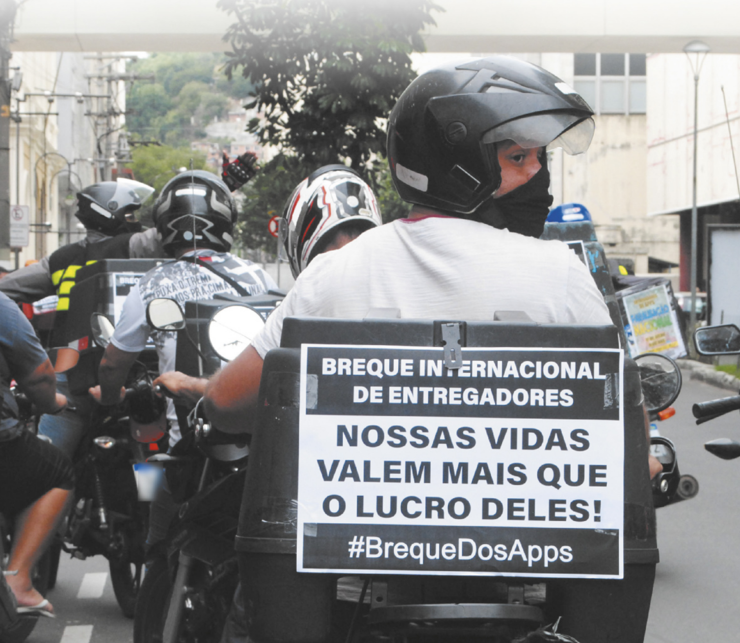 Fotografia de motociclistas dirigindo suas motos. No baú de uma moto tem um cartaz escrito 'Breque internacional de entregadores. Nossas vidas valem mais que o lucro deles! #BrequeDosApps'.