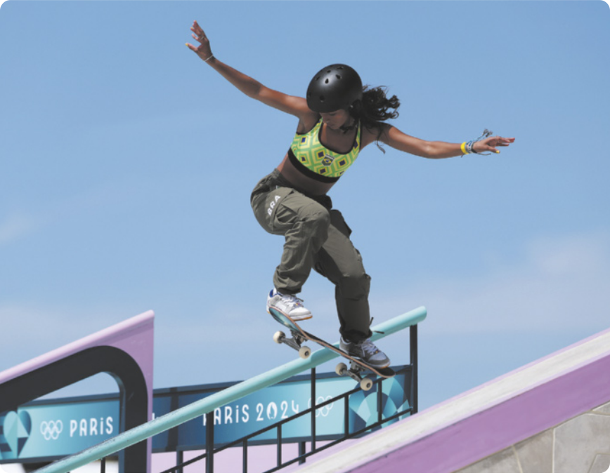 Fotografia de Rayssa Leal: uma garota de capacete com os braços abertos ao lado da cabeça e os joelhos flexionados, com os pés sobre o skate, executando uma manobra em que ela salta e desliza o skate sobre um corrimão.