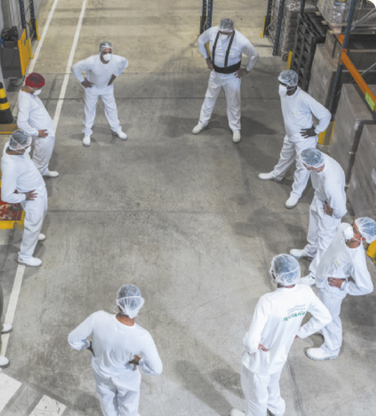 Fotografia de um grupo de pessoas com uniformes brancos, máscaras de proteção facial e toucas nos cabelos, em círculo, com as mãos na cintura em uma fábrica. Elas estão em pé girando os quadris, inclinando o tronco enquanto realizam o giro.