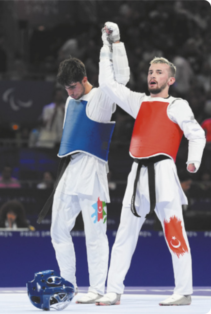 Fotografia de dois lutadores de taekwondo lado a lado em um ginásio. Eles estão usando coletes e equipamentos de proteção, segurando uma das mãos do adversário, erguida para o alto. Eles são Ali Can Özcan, à direita, com barba e cabelos curtos escuros; e Sabir Zeynalov, à esquerda, com barba e cabelos curtos e louros.