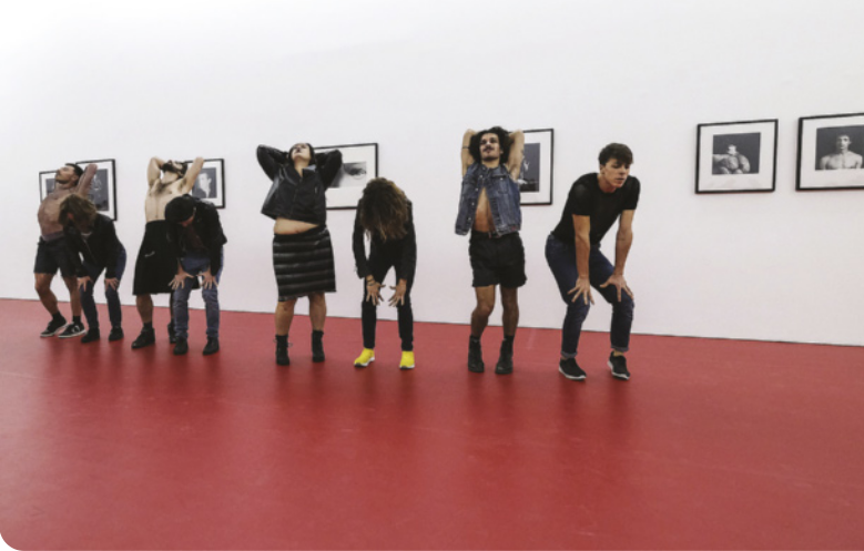 Fotografia de um grupo de pessoas enfileiradas em um espaço expositivo, dançando em pé em diversas posições.