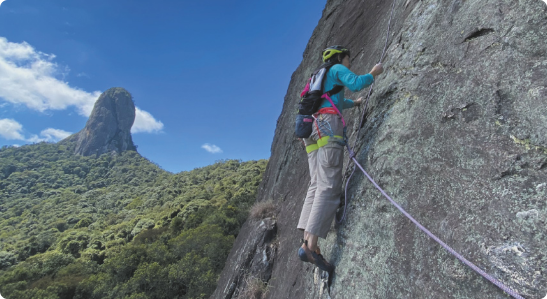 Fotografia de uma mulher escalando uma rocha em uma paisagem montanhosa, com a mata ao fundo. Ela usa capacete e outros equipamentos de escalada, com cordas de segurança presas aos equipamentos.