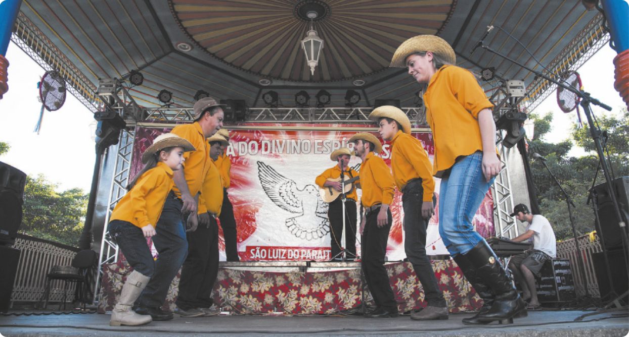 Fotografia de um grupo de homens, uma mulher e uma menina dançando frente a frente em um palco, usando chapéus e uniformizados com camisas amarelas. Eles estão organizados em duas fileiras, batendo com os pés no chão. Ao fundo do palco há um homem tocando violão diante de um microfone.
