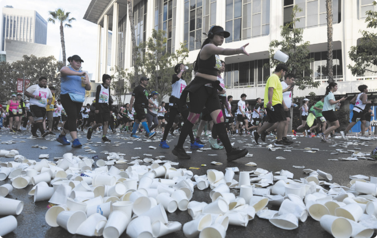 Fotografia de pessoas caminhando e correndo em uma maratona em uma via pública de Los Angeles. No chão há muitos copos de papel descartados e amassados, e uma mulher está arremessando um copo no chão.