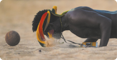 Fotografia de um homem indígena com o corpo deitado próximo ao chão, na areia, apoiado com as mãos, direcionando a cabeça a uma bola.