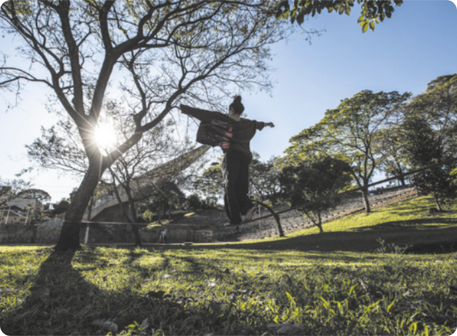 Fotografia de uma pessoa andando de braços abertos sobre uma corda bamba pendurada na horizontal, entre dois troncos de árvores, próxima ao chão de um gramado.