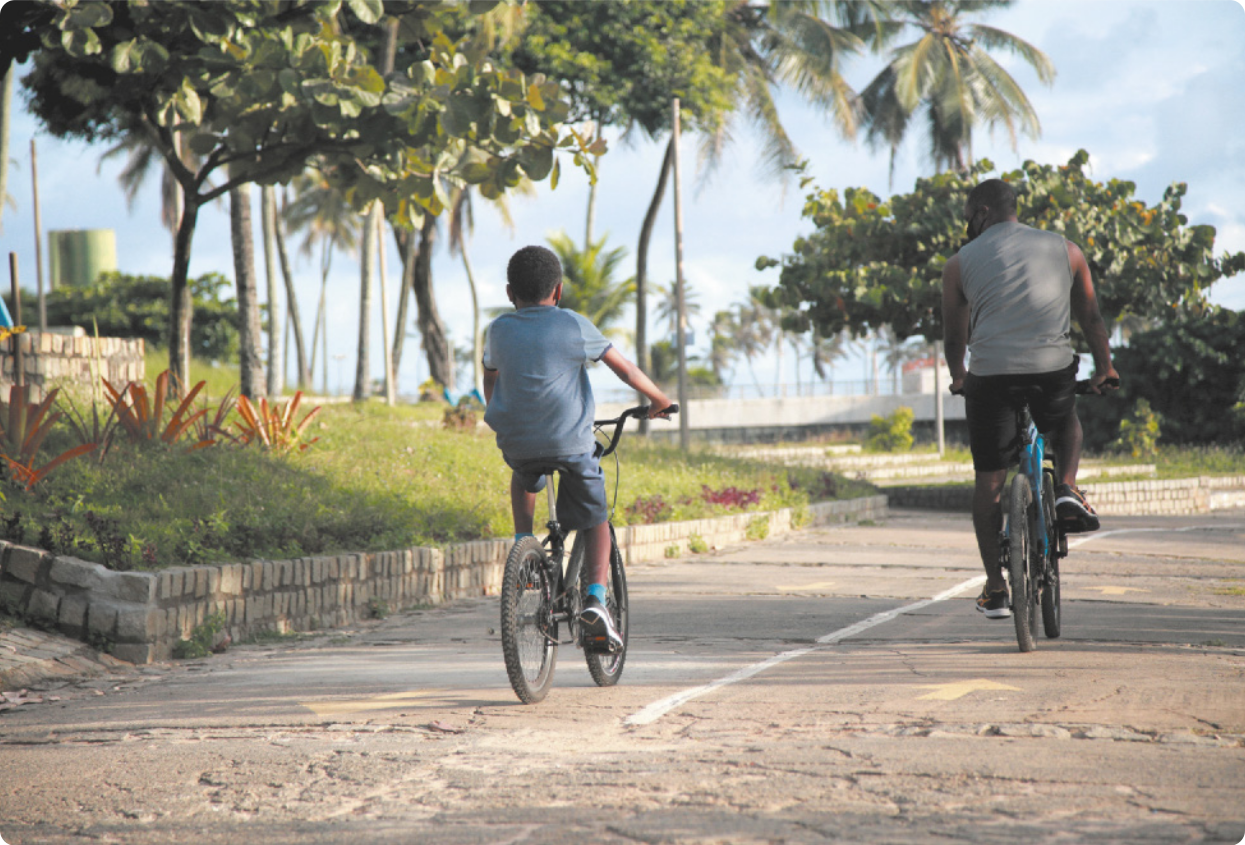 Fotografia de um menino e um homem andando de bicicleta lado a lado, em uma ciclovia de um parque.