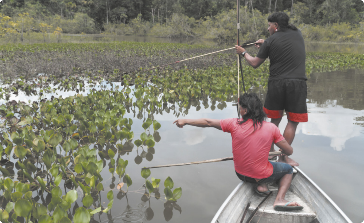 Fotografia de dois homens indígenas em um pequeno barco flutuando em um rio. Um deles está em pé, apontando um arco e flecha em direção à água; e o outro está sentado ao lado dele, segurando uma vara e apontando em direção à água.