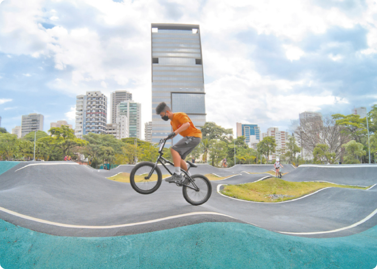 Fotografia de um homem andando de bicicleta em uma pista com relevo ondulado, com árvores e edifícios de uma cidade ao fundo.